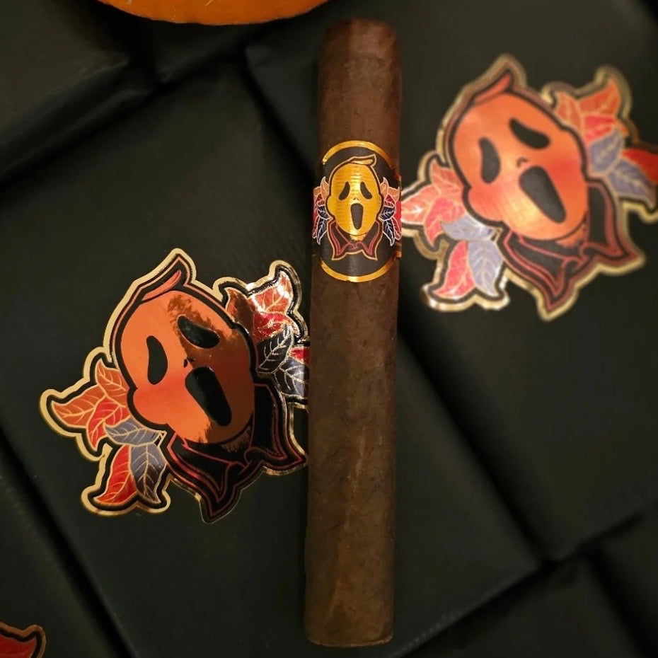 A single dark brown cigar with a Ghostface band, displayed on a black textured surface next to two Ghostface stickers and a pumpkin.